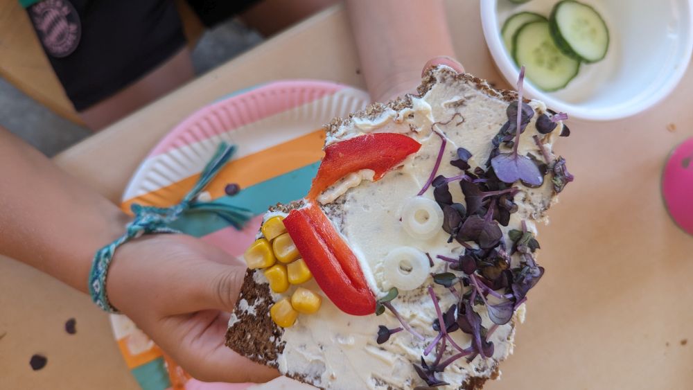 Hände halten ein Vollkornbrot mit Frischkäse und einem Gesicht aus Gemüse