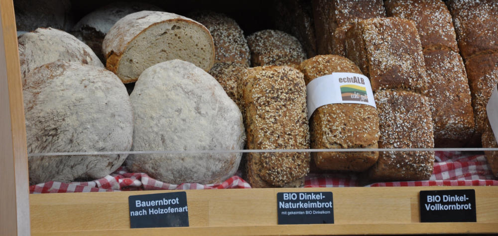3 verschiedene Brotsorten in einem Bäckerei-Regal. Beschriftung auf schwarzen Schildchen am Regal: Bauernbort nach Holzofenart, Bio-Dinkel-Naturkeimbrot und Bio-Dinkel-Vollkornbrot