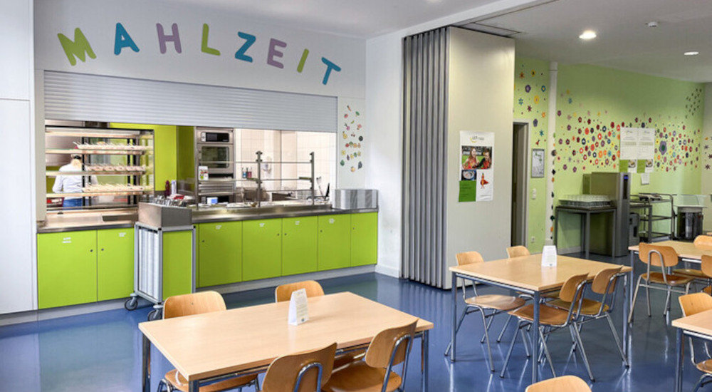 Blick in eine Schulmensa. Mehrere Tische mit jeweils 4 Stühlen aus hellem Holz. Die Mensa ist lichtdurchflutet. Im Hintergrund eine Ausgabetheke über der in bunten Buchstaben das Wort Mahlzeit steht. Die Farbe der Theke ist ein frisches Grün, eine Wand ist ebenfalls grün gestrichen und mit vielen kleinen Papierblumen geschmückt.