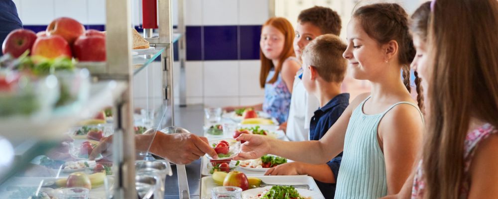 5 Schulkinder stehen mit gefüllten Tabletts an der Ausgabetheke in einer Schulmensa. Ein Mädchen nimmt lächelnd eine Schale mit Gemüse vom Ausgabepersonal entgegen.