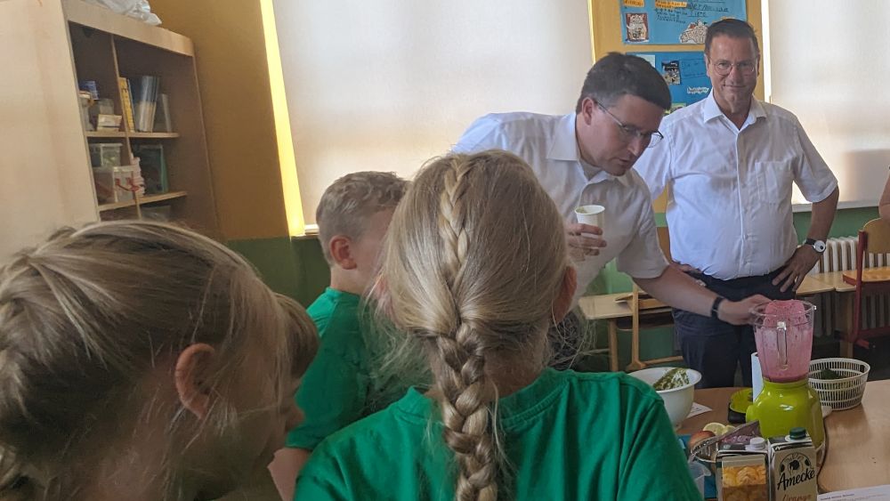 Kinder und Erwachsene stehen um einen Schultisch, auf dem ein Mixer mit einem roten Smoothie steht