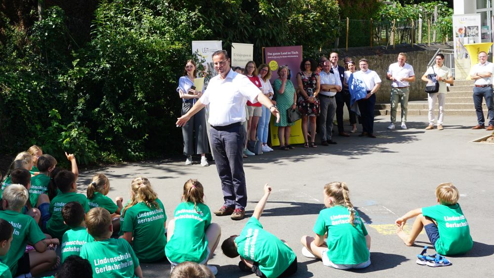 Sitzende Kinder und stehende erwachsene Gäste hören Minister Hauk MdL, bei seiner Ansprache zu 